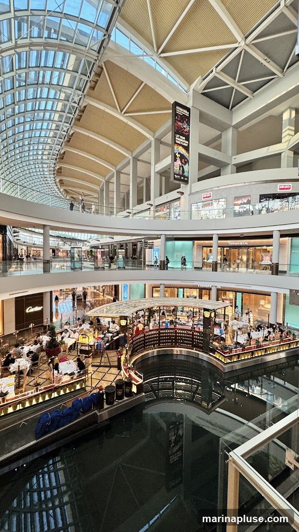 An interior view of a large, modern shopping mall. The architecture features a curved glass ceiling and multiple levels of shops and walkways. In the center, there is a canal with a circular dining area or bar overlooking it. People are seen dining and walking around. Brands like Cartier and Tiffany & Co. are visible on storefronts. A large vertical banner hangs from the ceiling, advertising an event or attraction.