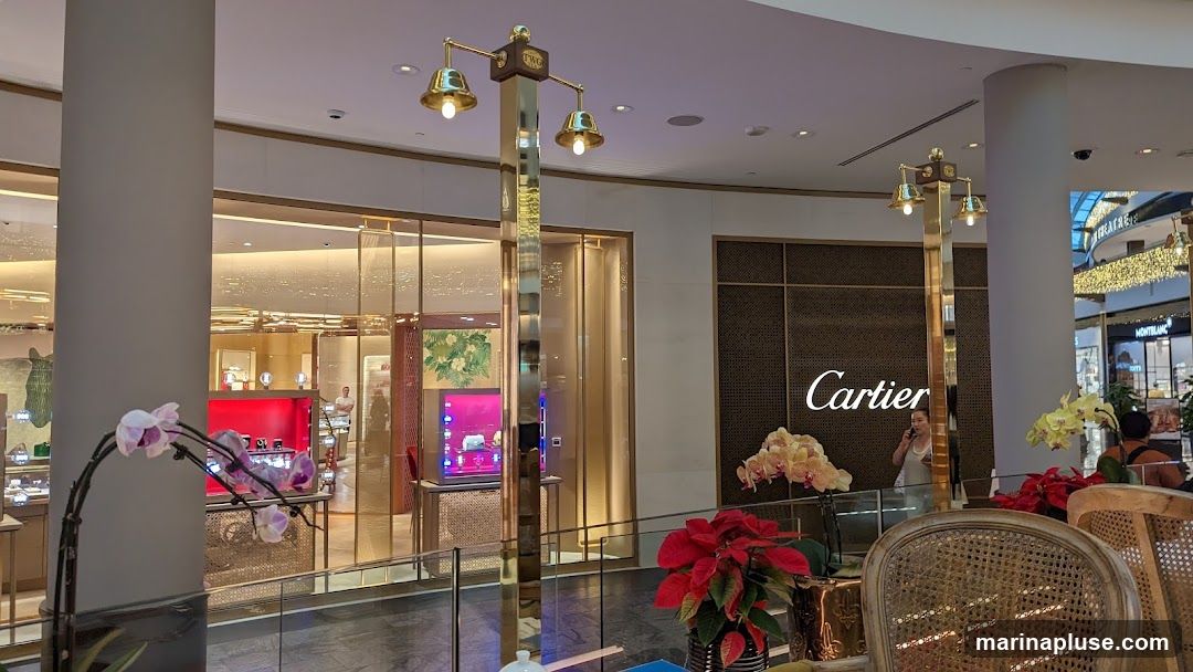 A view of the interior of a Cartier store, with displays of watches and jewelry. The Cartier logo is visible on a dark textured wall. In the foreground, there are decorative plants and seating areas. Other stores, including Montblanc, are visible in the background through the open mall area.
