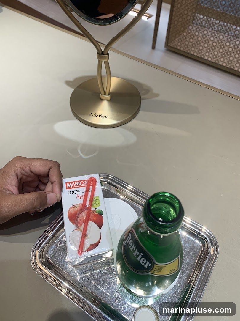 A close-up view of a silver tray holding a small carton of Marigo 100% Juice Apple drink with a red straw, and a partially consumed bottle of Perrier sparkling water. A hand is visible reaching for the juice box. In the background, a gold-toned vanity mirror with a circular base is prominently displayed, featuring the engraved word "Cartier". The setting appears to be an interior space, possibly a lounge or dressing area within a luxury establishment.