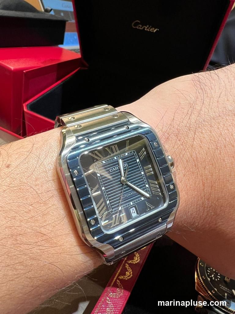 A close-up shot of a person's wrist wearing a silver Cartier watch. The watch face is dark with Roman numerals and the word "AUTOMATIC" is visible. In the background, a red Cartier box is open, revealing a black interior with the "Cartier" logo printed in gold. Another watch, partially visible, is in the lower right corner.
