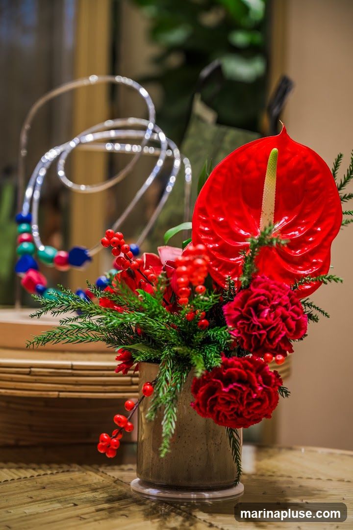 A close-up shot of a vibrant floral arrangement featuring a red anthurium flower, red carnations, and greenery, accented with red berries. The arrangement is in a textured ceramic vase. In the blurred background, a multi-colored beaded necklace is displayed on a stand. The arrangement sits on a woven surface.