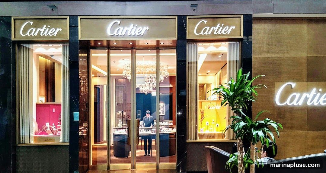 The exterior of a Cartier store is shown, featuring large display windows showcasing jewelry and watches. The store's name, "Cartier," is prominently displayed in illuminated lettering above the entrance and on the facade. A man in a suit stands inside near the entrance, attending to displays. Chandeliers are visible inside the store. Plants are placed outside the entrance.