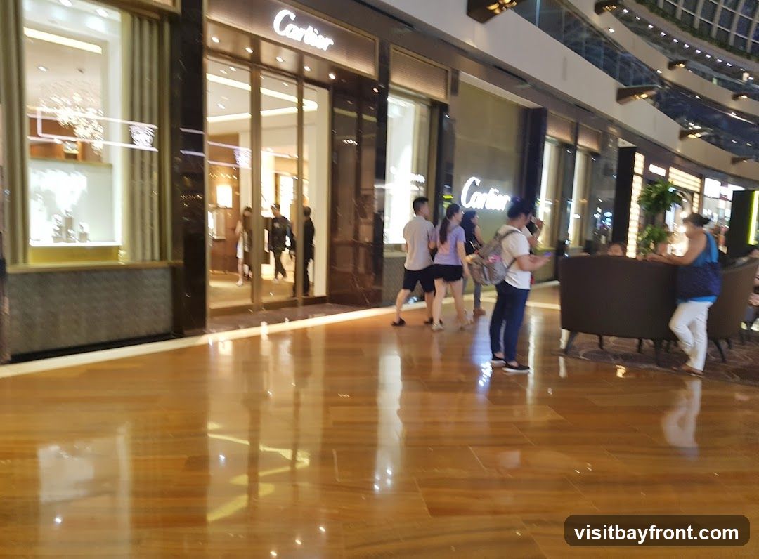 A wide shot of the exterior of a Cartier store in a mall. The store has large glass doors and windows, with the Cartier logo visible above the entrance. Several people are walking by the store. The floor is made of polished wood.