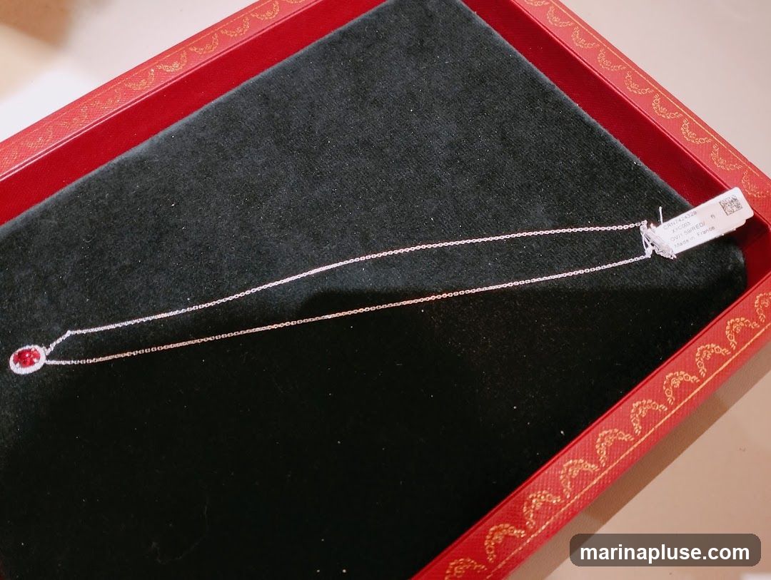 A delicate white gold necklace with a central oval ruby surrounded by small diamonds is displayed on a black velvet surface within a red, gold-embossed jewelry box. A white tag with product information is attached to the necklace chain.