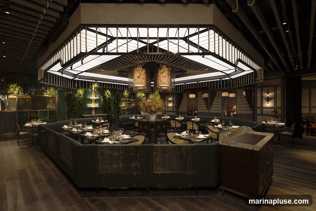 A well-lit, upscale restaurant interior with a modern Asian-inspired design. The dining area features plush green banquette seating arranged around tables set with white tableware. A central octagonal ceiling fixture with decorative lamps hangs above a large floral arrangement on a dark, ornate table. Green tiled walls and shelves with decorative items add to the ambiance. The flooring is dark wood.