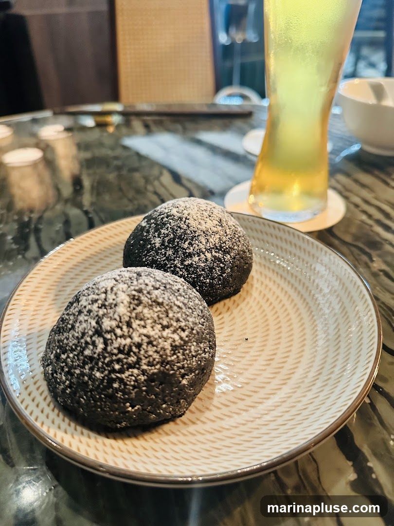 Two dark, round buns dusted with powdered sugar sit on a patterned plate. A tall glass of a light-colored beverage, possibly tea or a cocktail, is visible in the background, along with glimpses of a restaurant interior.