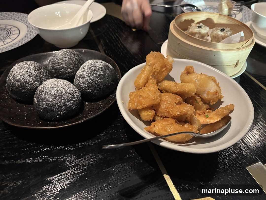 A close-up view of a table laden with various dim sum dishes. In the foreground, a dark plate holds four black, round buns dusted with powdered sugar. To the right, a white bowl contains a generous portion of golden-brown fried food, possibly fish or chicken, with a spoon resting inside. In the background, a bamboo steamer basket holds several pieces of dim sum, likely siu mai, topped with black dots. A white bowl and a plate with a blue and white pattern are also visible on the dark, glossy table.