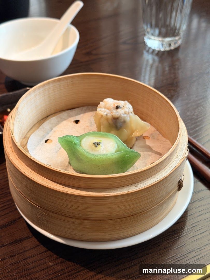 A bamboo steamer basket filled with two pieces of dim sum sits on a white plate on a wooden table. One piece is a green dumpling shaped like a pea pod with a white filling and black seeds. The other is a light yellow dumpling with a textured top. A white ceramic spoon rests in a white bowl in the background, and chopsticks are visible to the right. A glass of water and a small dish of sauce are also present.