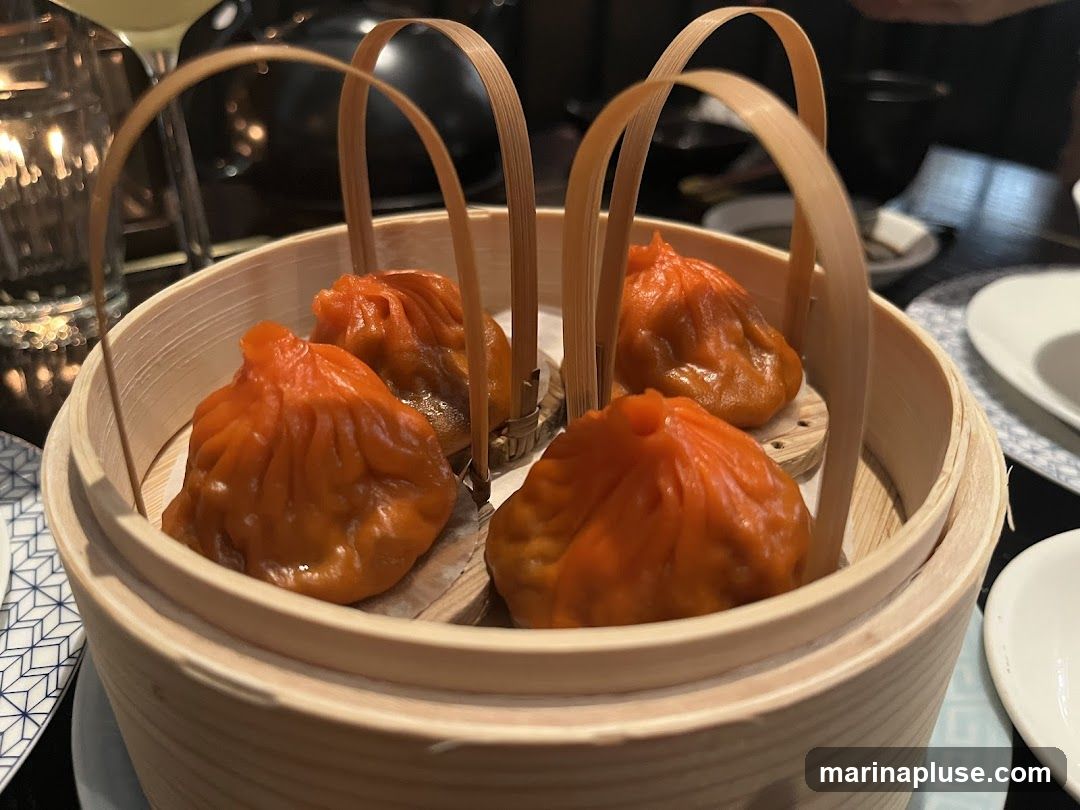 A close-up shot of four orange-colored steamed dumplings arranged in a bamboo steamer. The dumplings have a pleated top and are sitting on small wooden discs within the steamer. The steamer has two curved bamboo handles. In the background, a dimly lit restaurant setting is visible with a glass, a candle, and plates.