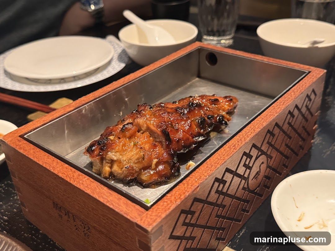 A close-up shot of a glazed meat dish served in a wooden box with a metal insert. The meat appears to be roasted or grilled and is coated in a dark, glossy sauce. The wooden box has intricate geometric carvings and a circular emblem with a bird silhouette. Several small white bowls and plates are visible in the background, along with chopsticks and a glass of water. The setting suggests a restaurant environment.