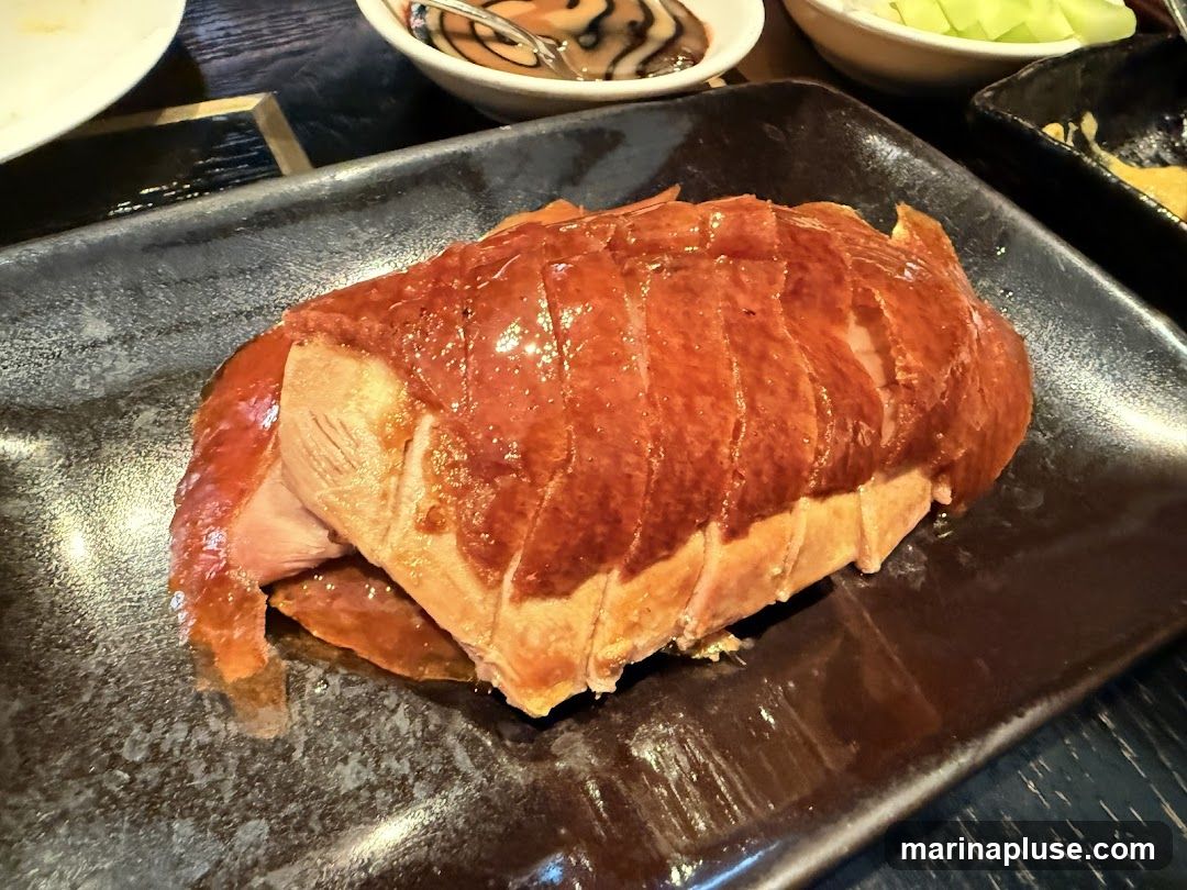 A close-up shot of a roasted Peking duck, glistening with a rich, reddish-brown glaze. The duck is carved into thick slices, revealing tender meat beneath the crispy skin. It is served on a dark, textured rectangular plate, with glimpses of other dishes and bowls in the background.