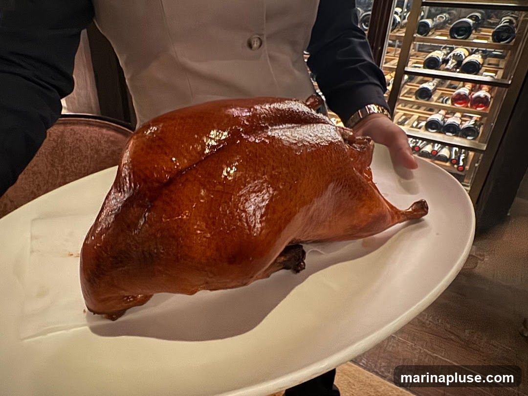 A whole roasted duck with crispy, golden-brown skin is presented on a white platter by a server. In the background, a wine rack filled with bottles is visible.
