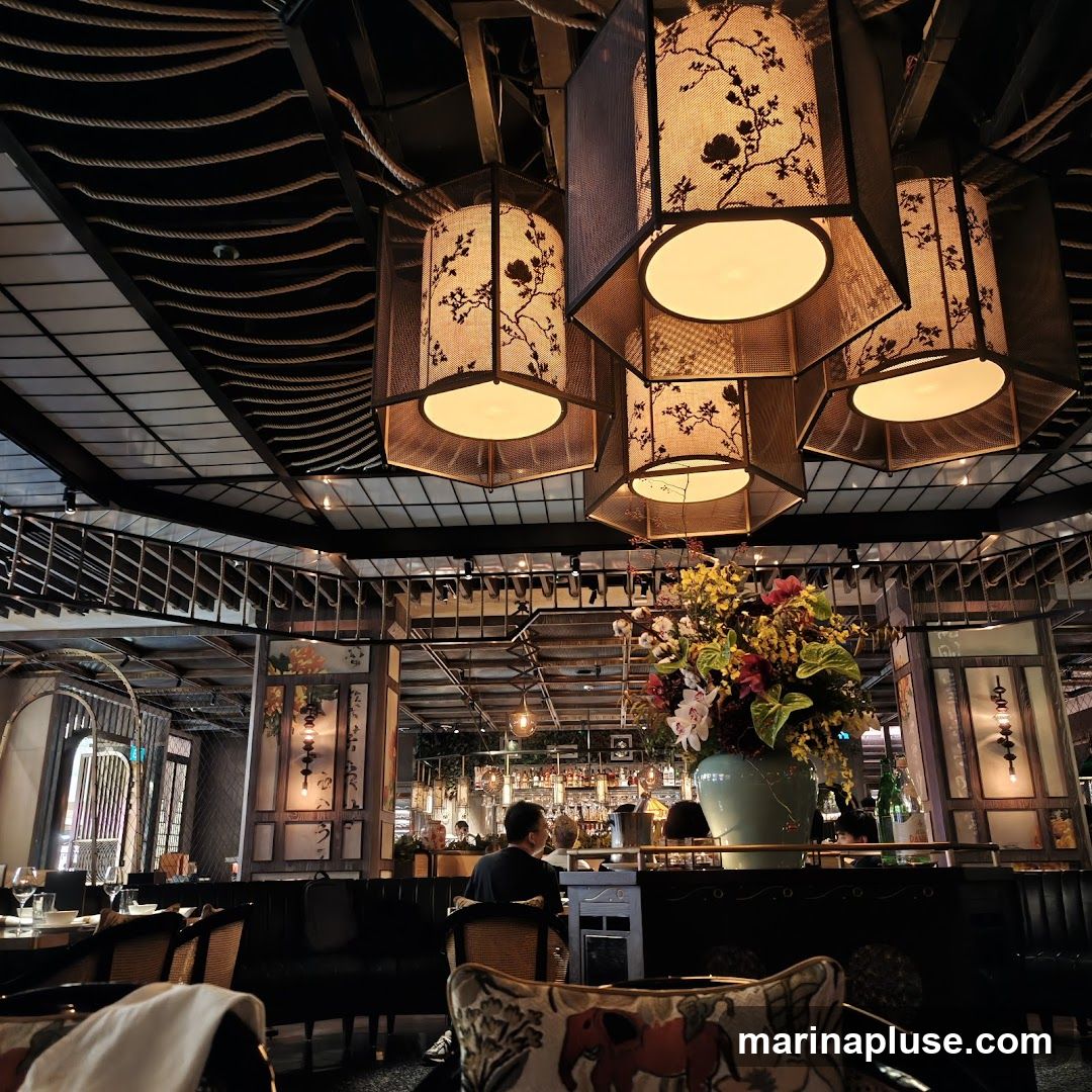 The image shows the interior of a restaurant with a focus on the ceiling and decorative elements. Several large, hexagonal lanterns with floral patterns hang from the ceiling, casting a warm light. A large vase filled with colorful flowers is prominently displayed in the foreground. In the background, a bar area is visible with patrons seated. The overall ambiance appears sophisticated and dimly lit.