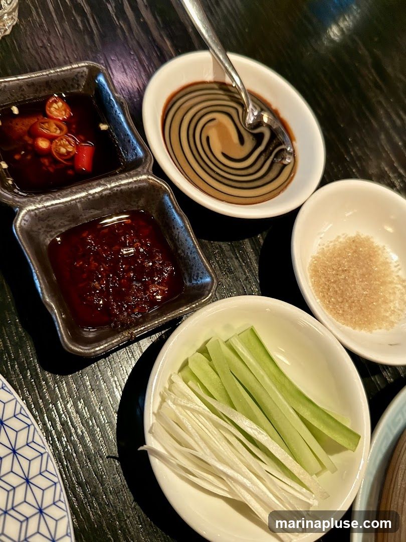 A close-up view of various condiments and garnishes arranged on a dark wooden table. In the foreground, a white bowl contains julienned scallions and cucumber sticks. To the left, a dark, multi-compartment dish holds two types of sauces: one with visible chili slices and another darker, thicker sauce. A separate white oval dish contains a swirling pattern of two sauces, with a spoon resting in it. Another small white bowl holds a pile of granulated sugar.