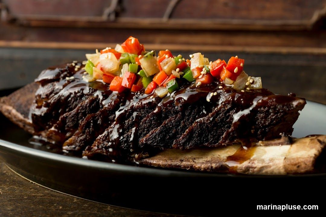 A close-up shot of braised short ribs glazed in a dark sauce and topped with a finely diced mixture of red and green bell peppers, onions, and sesame seeds. The ribs are served on a dark plate.