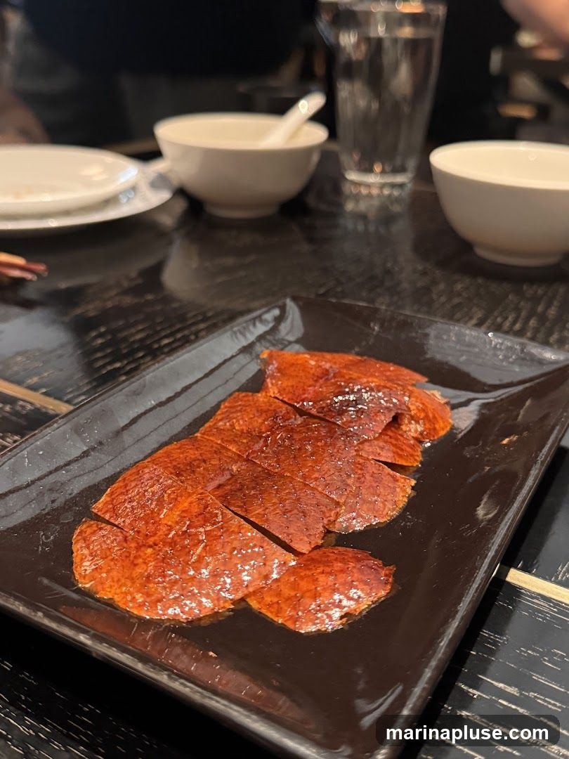A close-up shot of crispy Peking duck skin served on a black rectangular plate. The skin is glistening and has been sliced into pieces. In the background, there are bowls, a glass of water, and a plate, suggesting a dining setting.