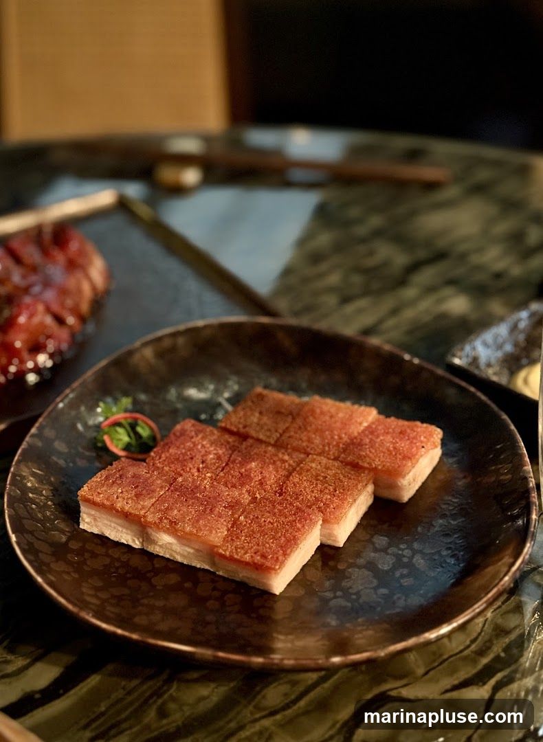 Two plates of food are visible on a dark, textured table. The plate in the foreground contains several rectangular slices of crispy pork belly with a golden-brown, crackled skin and a layer of white fat and meat. A small garnish of green herbs and red slivers is placed on the side. In the background, another plate holds slices of glazed, dark red meat, possibly char siu. Chopsticks are visible in the background.
