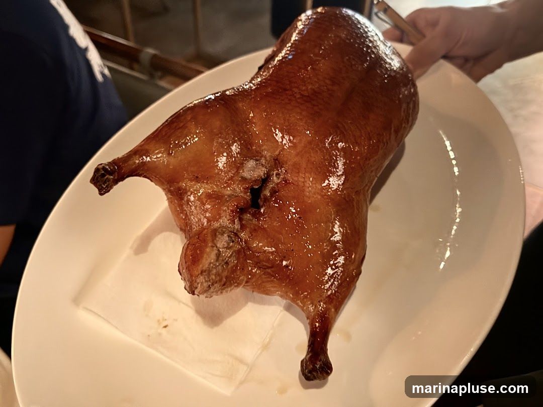 A whole roasted duck with crispy, glossy skin is presented on a white oval platter. The duck is positioned on a white napkin. A hand is visible in the upper right corner, holding a utensil near the duck's head. Another person is partially visible on the left side of the image.