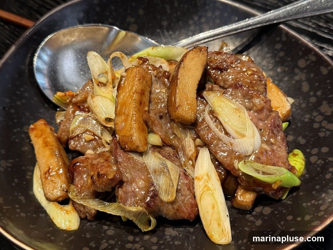 A close-up shot of a stir-fry dish featuring thinly sliced beef, pieces of mushroom, and scallions in a glossy sauce. The dish is served in a dark, textured bowl with a spoon and fork resting on the side.