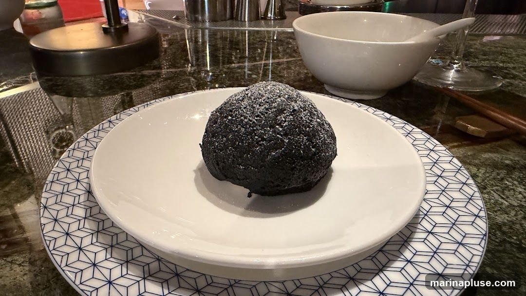 A dark, spherical dessert dusted with powdered sugar sits on a white plate with a blue geometric pattern. The plate is on a dark, reflective surface, possibly a marble tabletop. In the background, a white bowl, chopsticks, and part of a lamp are visible.