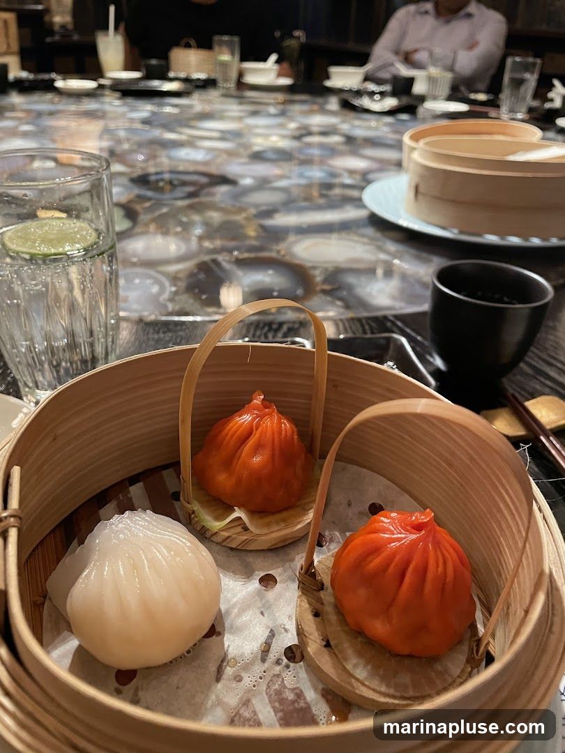 A close-up view of a bamboo steamer basket filled with dim sum. There are three dumplings visible: one white, translucent dumpling and two bright orange, soup-filled dumplings. The dumplings are served in individual bamboo cups. In the background, a table is set for a meal with drinks and other diners. The table has a unique, agate-like patterned surface.