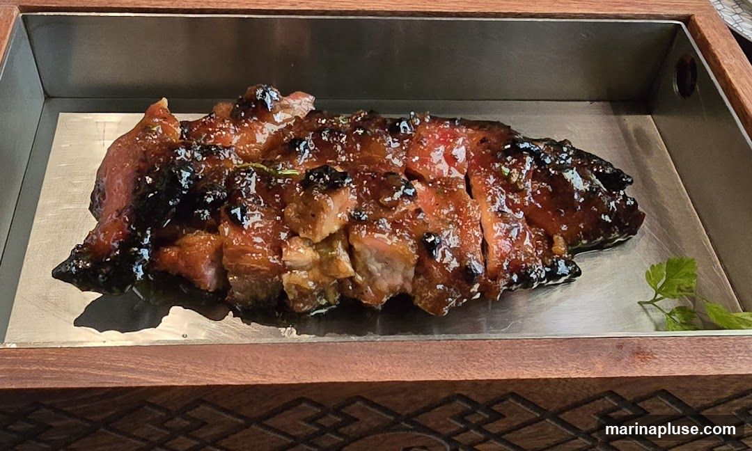 A close-up shot of sliced char siu pork, a Chinese barbecue pork dish, served in a metal tray with a wooden border. The pork is glazed with a dark, glossy sauce and appears to be garnished with herbs.