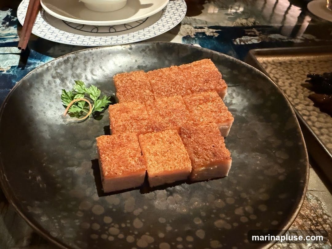 A close-up shot of several rectangular slices of crispy pork belly, arranged on a dark, textured plate. Each slice has a golden-brown, crackling exterior and a visible layer of fat and meat. A small sprig of green parsley garnishes the plate. In the background, a stack of plates and chopsticks are visible, along with a glimpse of a patterned tablecloth. Another dish with dark food items is partially visible to the right.
