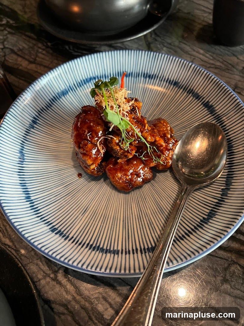A close-up shot of a plate of glazed appetizers, possibly chicken wings or similar, served on a blue and white striped ceramic plate. The food is coated in a dark, glossy sauce and garnished with microgreens and crispy threads. A silver spoon rests on the right side of the plate. The table surface appears to be dark marble.