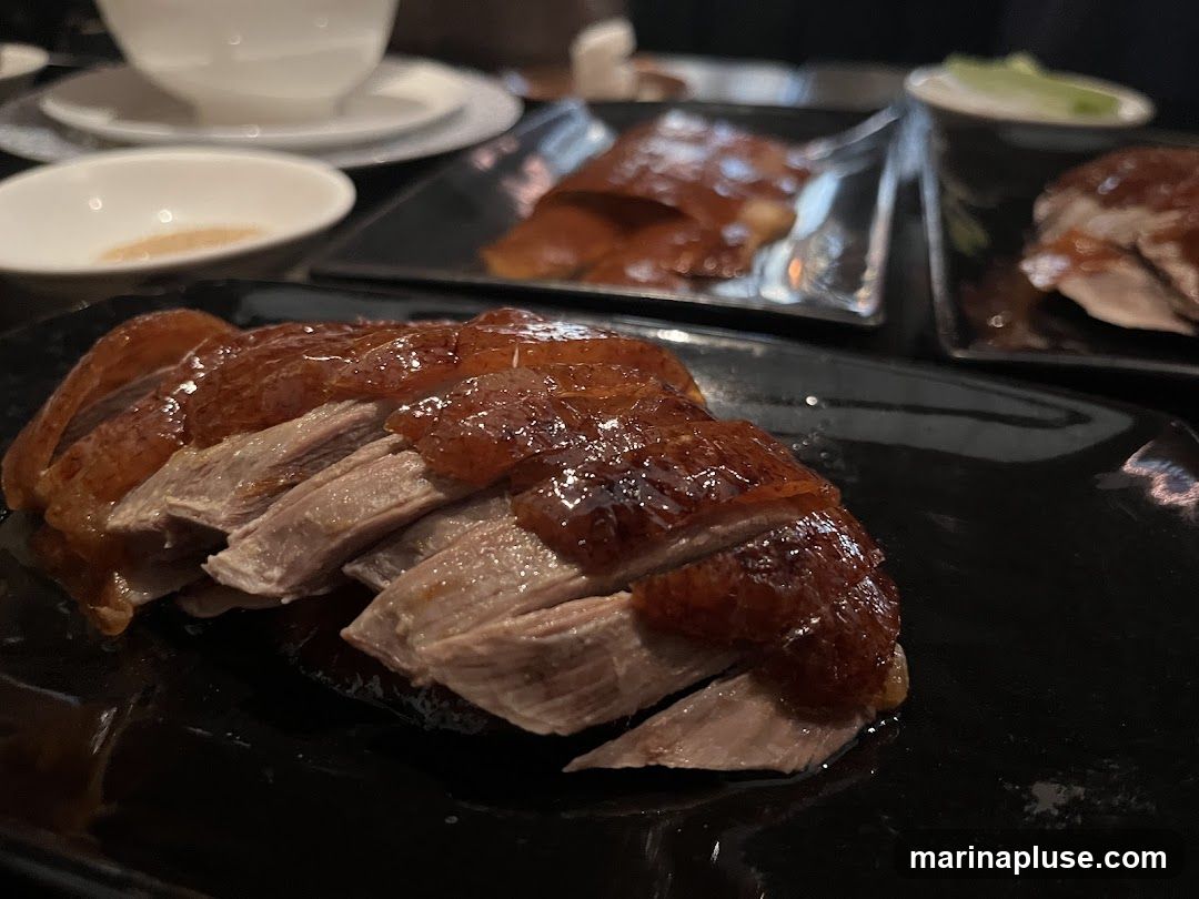 Slices of roasted Peking duck with crispy skin are served on black plates. In the background, other plates of duck and small dishes are visible, along with a white bowl and saucer.