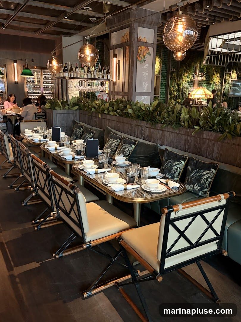 A view of a restaurant interior with a long banquette seating area and tables set for dining. The tables are adorned with white tableware, napkins, and small black menu holders. Behind the banquette, there is a wall decorated with botanical artwork and green tiled accents. A bar area with wine bottles and glassware is visible in the background, along with patrons seated at tables. The lighting is warm, with decorative pendant lamps and a table lamp.