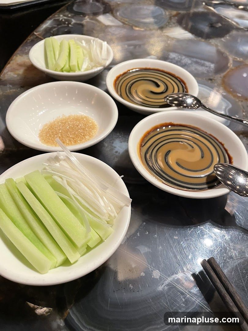 A close-up overhead view of several small white bowls containing condiments and sliced vegetables, likely for a meal. In the foreground, a bowl holds sliced cucumber and scallions. To its right, two bowls contain a dark sauce swirled with a lighter sauce, each with a spoon. Another small bowl holds granulated sugar. The table surface appears to be a dark, polished material with a marbled pattern. Chopsticks rest on the table.