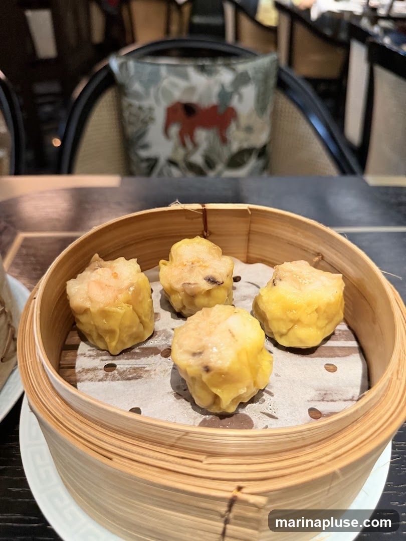 A close-up shot of four steamed dumplings, likely siu mai, served in a traditional bamboo steamer. The dumplings are golden yellow and appear to be filled with shrimp and possibly mushrooms. The steamer is placed on a white plate with a decorative rim. In the background, out of focus, are chairs with patterned cushions featuring a red camel-like animal. The setting suggests an indoor dining environment.