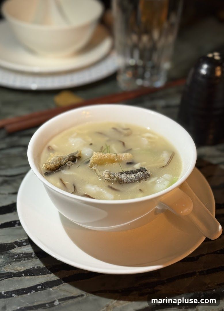 A close-up shot of a bowl of fish maw soup, garnished with crispy fish skin and herbs. The soup is creamy and appears to contain pieces of fish maw and other ingredients. The bowl is white and sits on a matching saucer. In the background, out of focus, are stacked bowls, chopsticks, a glass of water, and a salt shaker. The table has a dark, striped pattern.