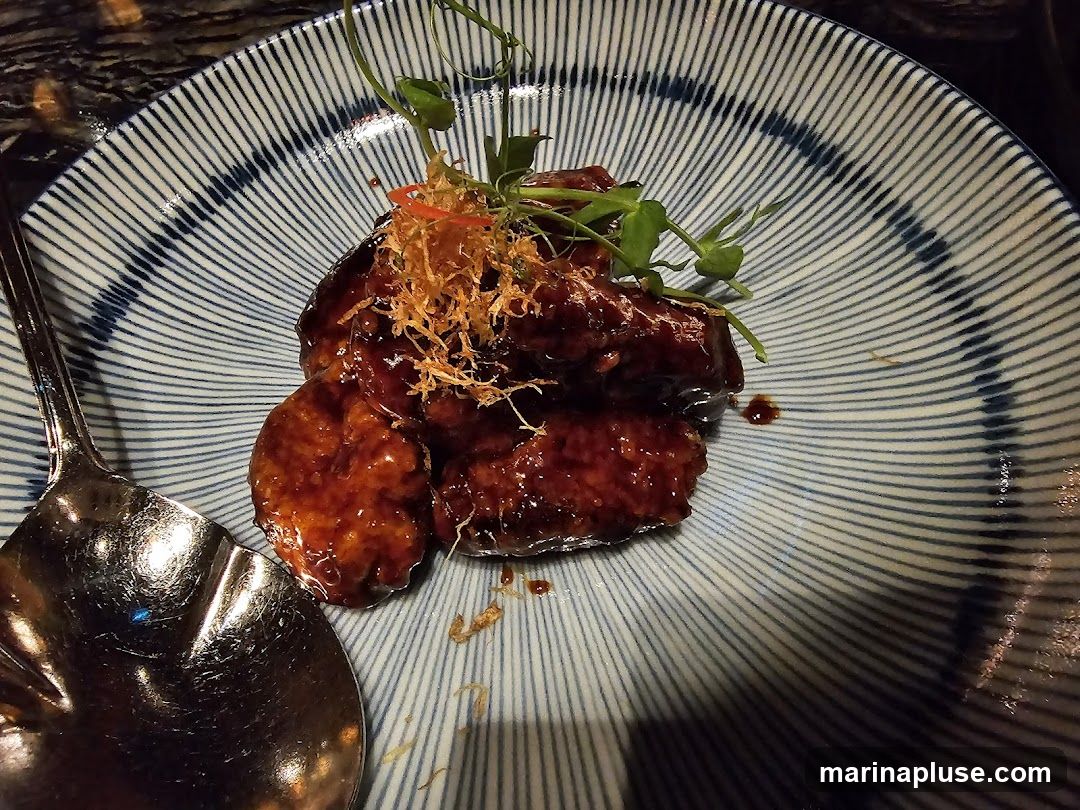 A close-up shot of a plate of glazed meat, possibly ribs or wings, served with a garnish of microgreens and crispy fried shallots. The dish is presented on a blue and white striped ceramic plate, with a large metal spoon to the left. The meat is coated in a dark, glossy sauce.