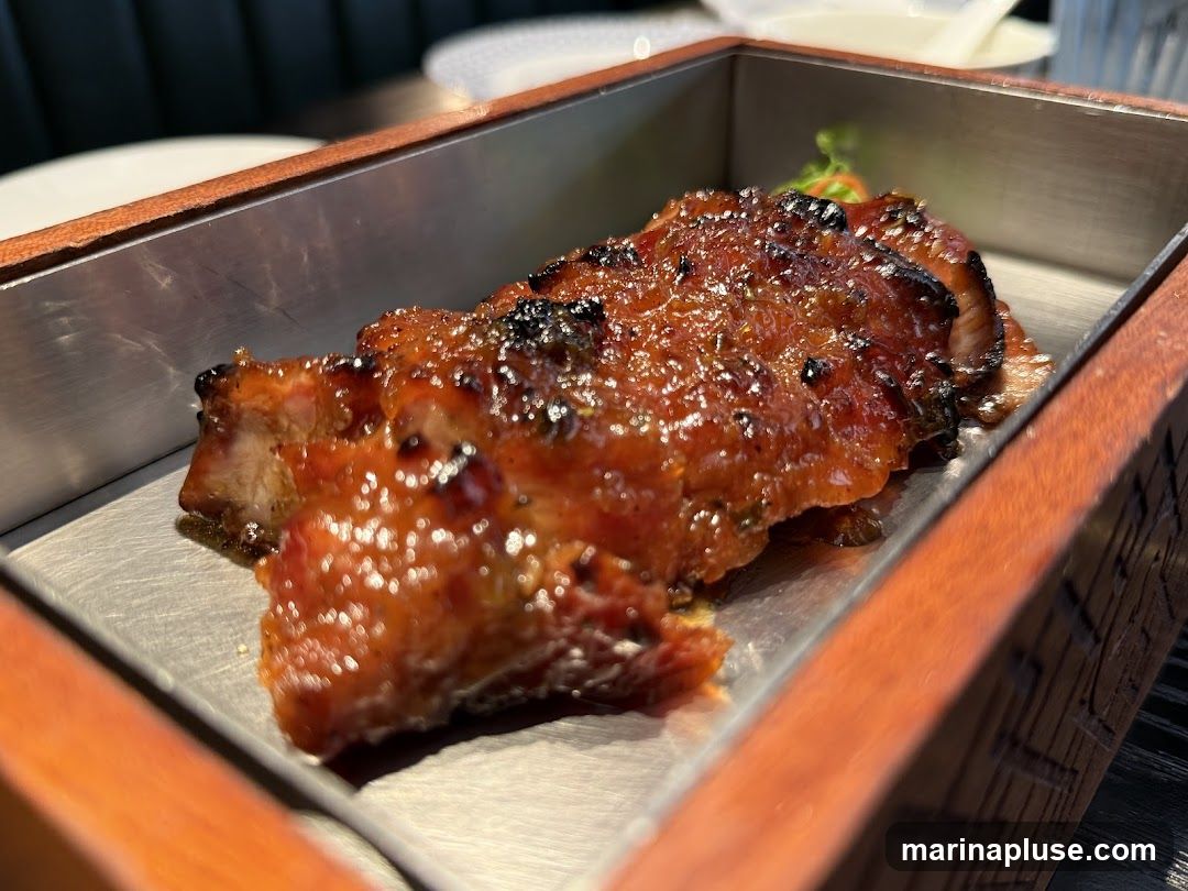A close-up shot of glistening char siu pork, a popular Chinese barbecued pork dish, served in a rectangular metal dish with wooden trim. The pork is glazed with a reddish-brown sauce and has some charred edges, indicating it has been roasted or grilled. A small garnish of green and orange shredded vegetables is visible at the back of the dish. The lighting highlights the glossy texture of the meat.