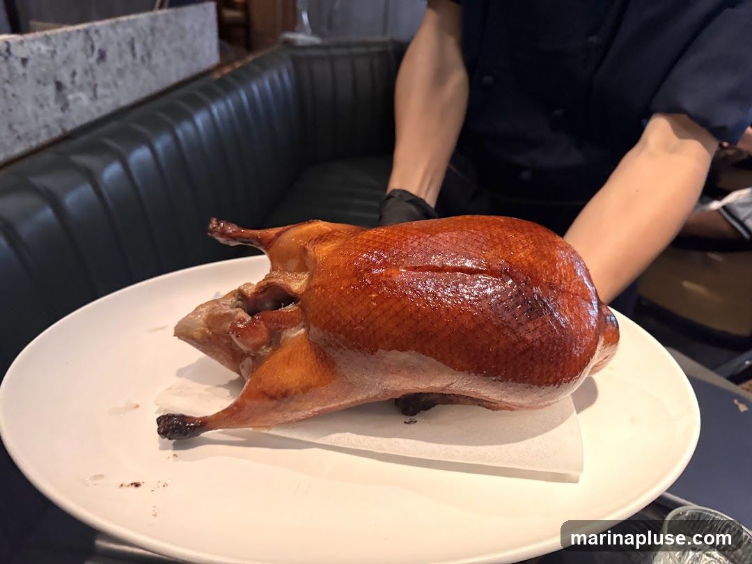 A whole roasted Peking duck is presented on a white plate, resting on a piece of white paper. The duck has a glossy, crispy, golden-brown skin. A person wearing a dark uniform and black gloves is holding the plate, serving the duck. The background shows a dark green tufted booth seating and a glimpse of a marble countertop.