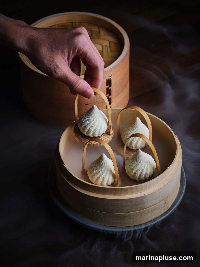 A hand is shown reaching for a small, white, textured dumpling served in a miniature bamboo basket. The basket is placed within a larger bamboo steamer, which is emitting a wispy vapor. Several other similar dumplings are visible in the steamer. The presentation is dramatic and artistic, with dark lighting and swirling smoke or vapor.