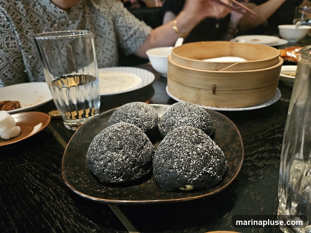 A close-up shot of four dark, round desserts dusted with powdered sugar, served on a black plate. In the background, a bamboo steamer basket sits on a table, along with a glass of water and other dishes. People are visible in the background, suggesting a dining setting.