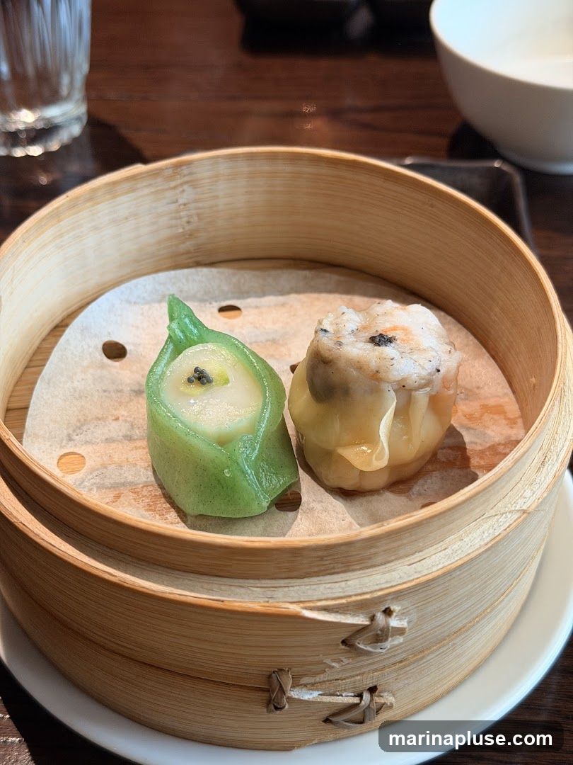 A close-up shot of two pieces of dim sum in a bamboo steamer basket. One is a green, translucent dumpling shaped like a leaf, filled with a white creamy substance and topped with black seeds. The other is a traditional siu mai, a steamed pork and shrimp dumpling, topped with a piece of shrimp and black pepper. The steamer basket is placed on a white plate, and the background shows a wooden table, a glass of water, and a white bowl.