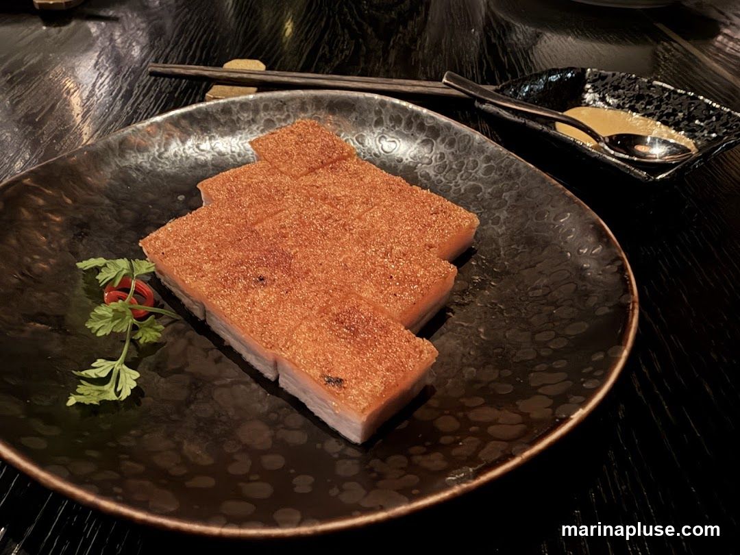 A close-up shot of crispy pork belly slices on a dark, textured plate. The pork has a golden-brown, crackling skin and is garnished with a sprig of parsley and a small red chili. A small dish with a spoon and a yellow sauce is visible in the background, along with chopsticks.