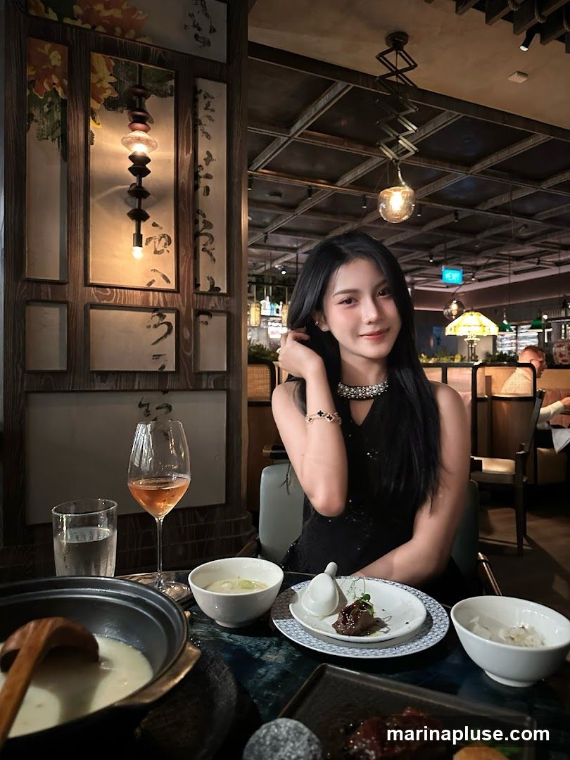 A woman in a black dress sits at a table in a restaurant, with a meal laid out before her. The table has a pot of soup, bowls of rice and other dishes, and a glass of rosé wine. The restaurant interior features decorative wooden panels with Asian-inspired artwork and modern lighting fixtures.