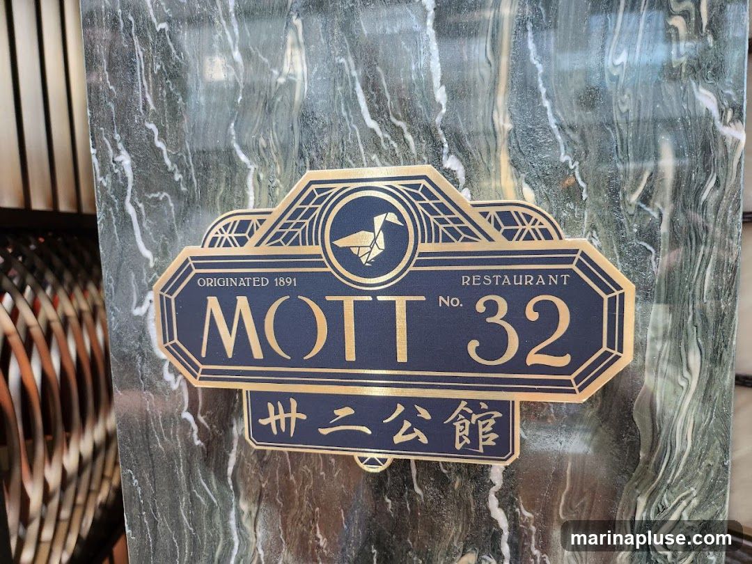 A close-up shot of a restaurant sign with a dark blue background and gold lettering. The sign reads "MOTT NO. 32 RESTAURANT ORIGINATED 1891" and also features Chinese characters below. The sign is mounted on a dark green marble wall. To the left, a blurred view of a decorative metal structure is visible.