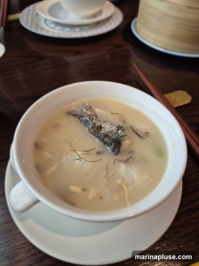 A close-up shot of a bowl of fish soup with a piece of fish skin on top. The soup is creamy and appears to have chunks of fish and other ingredients. Chopsticks and a bamboo steamer are visible in the background, suggesting an Asian dining setting.