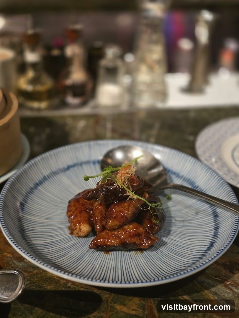 A close-up shot of a plate of glazed appetizers, possibly chicken wings or dumplings, served with a garnish of microgreens. The food is coated in a dark, glossy sauce and arranged on a blue and white striped plate. In the blurred background, bottles of condiments and other restaurant elements are visible.