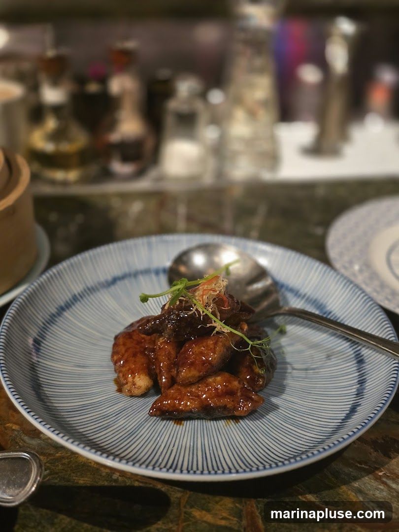 A close-up shot of a plate of glazed appetizers, possibly chicken wings or dumplings, served with a garnish of microgreens. The food is coated in a dark, glossy sauce and arranged on a blue and white striped plate. In the blurred background, bottles of condiments and other restaurant elements are visible.
