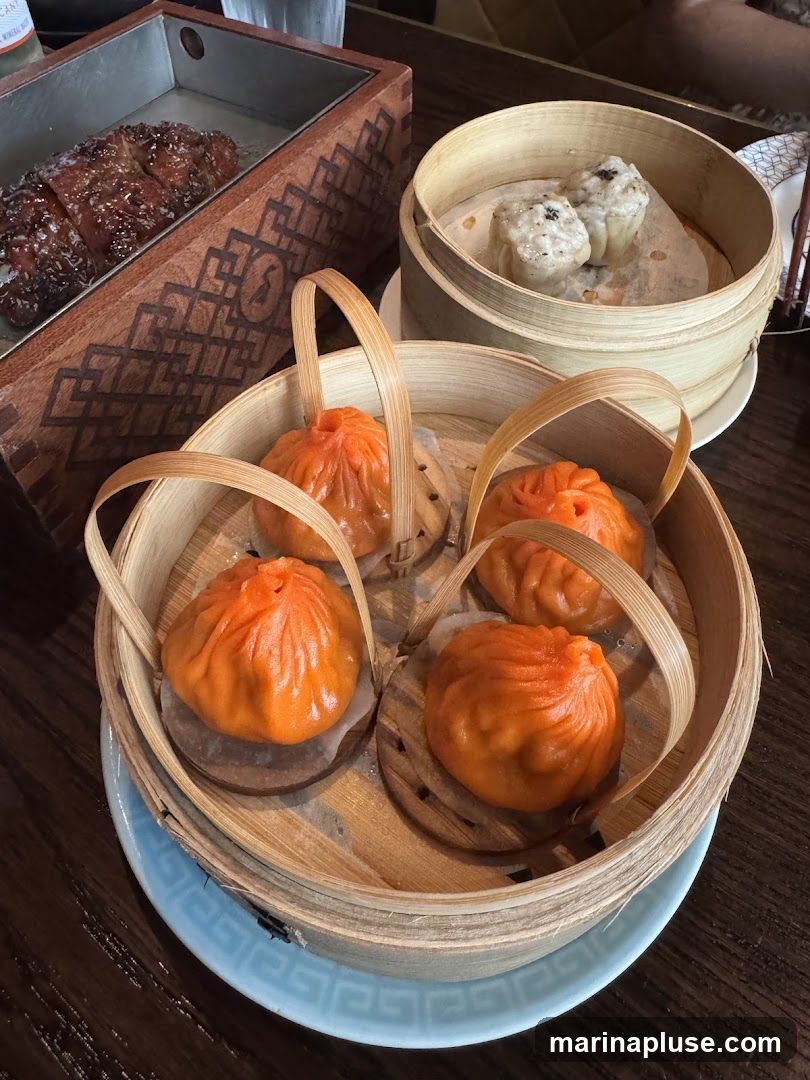 A close-up view of several bamboo steamers filled with food. In the foreground, a steamer contains four bright orange, soup-filled dumplings. Behind it, another steamer holds two dim sum dumplings. To the left, a rectangular metal tray with a wooden decorative border holds slices of glazed meat, possibly char siu. The food is arranged on a dark wooden table.