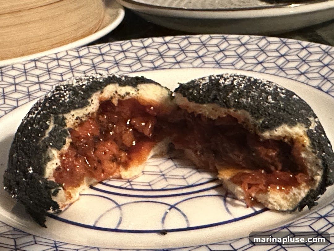 A close-up shot of a black sesame bun, split in half, revealing a glistening red filling. The bun has a soft, white interior and a dark, textured exterior coated in black sesame seeds and a dusting of powdered sugar. The filling appears to be a sweet or savory paste, possibly with small pieces of ingredients. The bun is served on a white plate with a blue geometric pattern. In the background, a bamboo steamer basket is visible.
