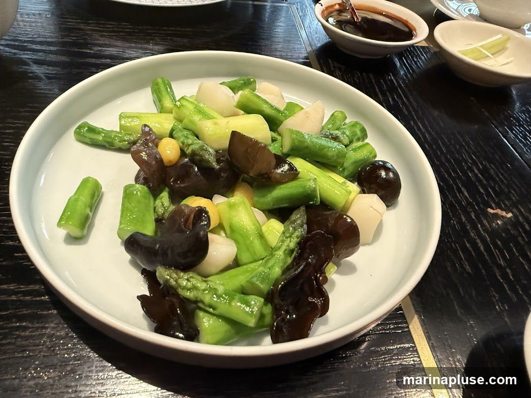 A close-up shot of a white plate filled with a stir-fry dish. The dish contains chopped asparagus spears, dark wood ear mushrooms, and what appear to be water chestnuts or similar white vegetables. A small dish of dark sauce and another with sliced green onions are visible in the background, along with a portion of a patterned plate. The food is served on a dark, textured wooden table.