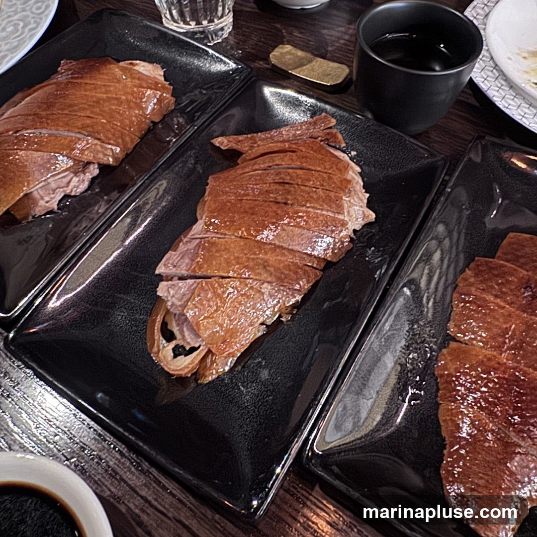 Three black rectangular plates are filled with sliced Peking duck. The duck skin is a deep golden brown and appears crispy. The meat underneath is moist and tender. The plates are arranged on a dark wooden table. A small black cup filled with a dark liquid and a small golden object are visible in the background.