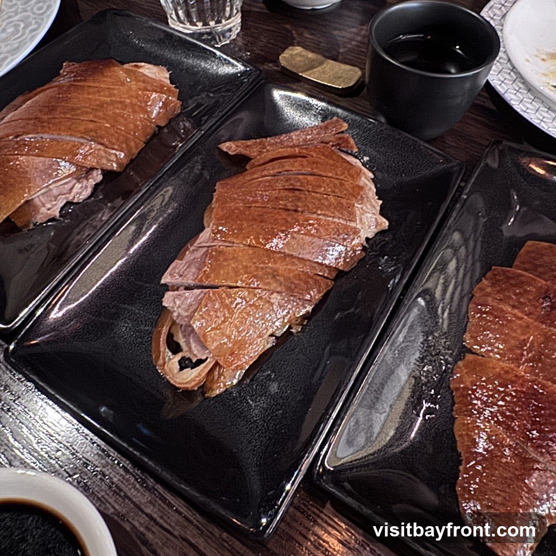 Three black rectangular plates are filled with sliced Peking duck. The duck skin is a deep golden brown and appears crispy. The meat underneath is moist and tender. The plates are arranged on a dark wooden table. A small black cup filled with a dark liquid and a small golden object are visible in the background.